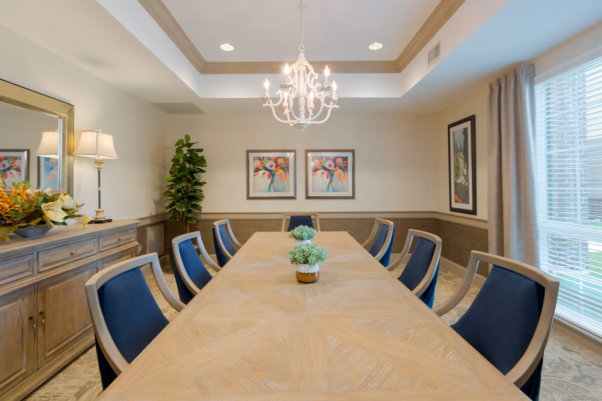 A well-lit dining room with a long wooden table surrounded by eight chairs with blue cushions. The room features a chandelier hanging from a tray ceiling, two colorful floral paintings on the wall, a large window with curtains, a wooden sideboard with a lamp and floral arrangement, and a tall green plant in the corner.