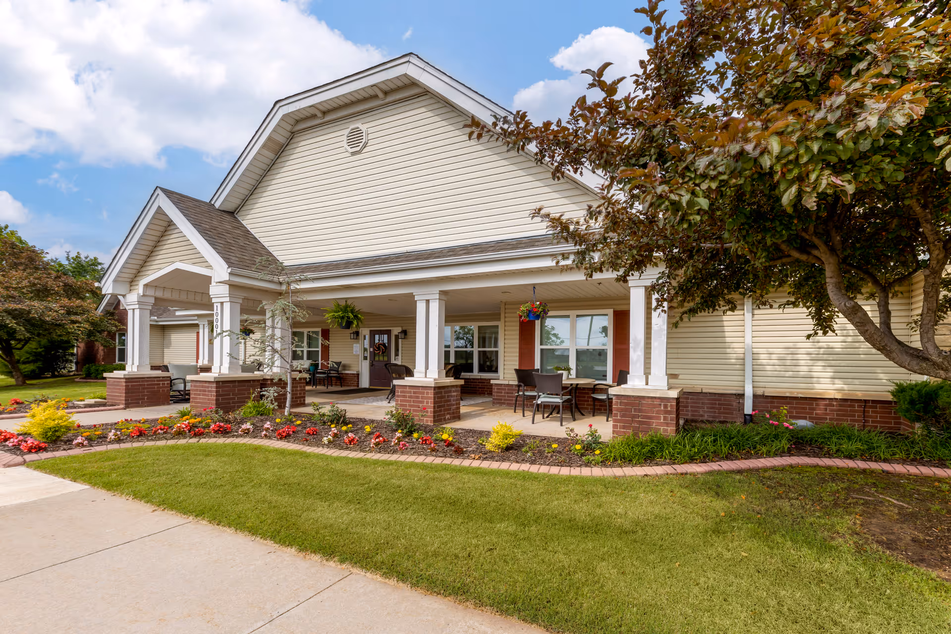 Front exterior view of a single-story building with beige siding and a gabled roof, featuring a covered porch with white columns and brick bases. The porch has outdoor seating and hanging flower baskets. The surrounding area includes a well-maintained lawn, flower beds with colorful flowers, and a tree on the right side under a partly cloudy sky.