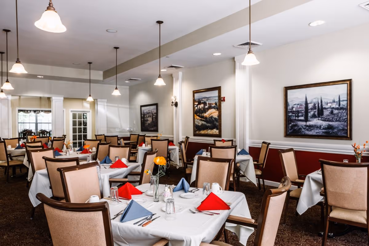 A dining room with multiple tables covered in white tablecloths, each set with red and blue folded napkins, plates, glasses, and silverware. The room features beige upholstered chairs, hanging pendant lights, and framed landscape paintings on the walls.