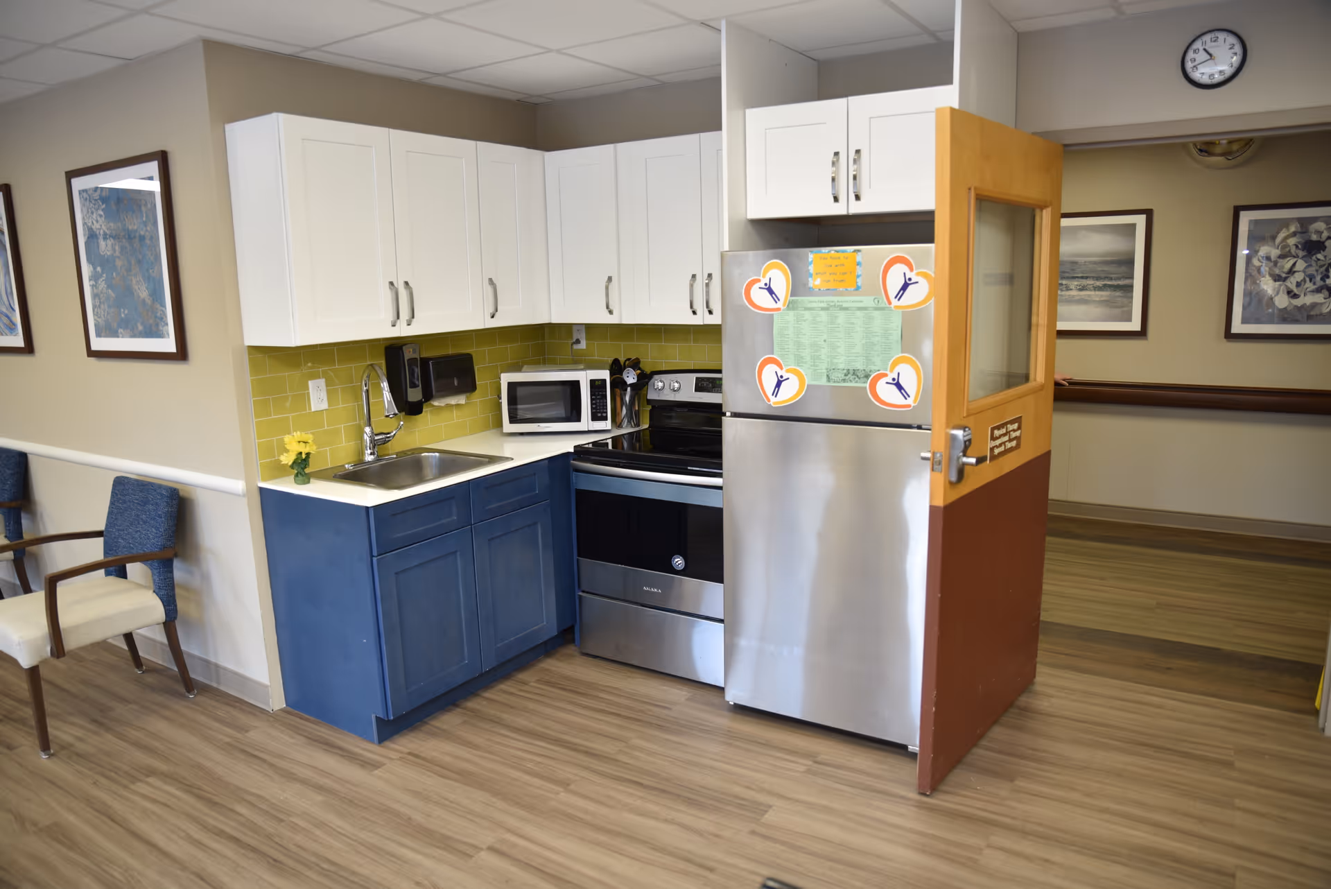 A small kitchen area in a senior living facility with white upper cabinets and blue lower cabinets. The kitchen includes a stainless steel refrigerator with magnets and notes, a microwave, an electric stove, and a sink with a faucet. The walls are beige with framed artwork, and there are two chairs with blue and beige upholstery to the left. The floor is wood-patterned, and a clock is visible on the wall above the open door.