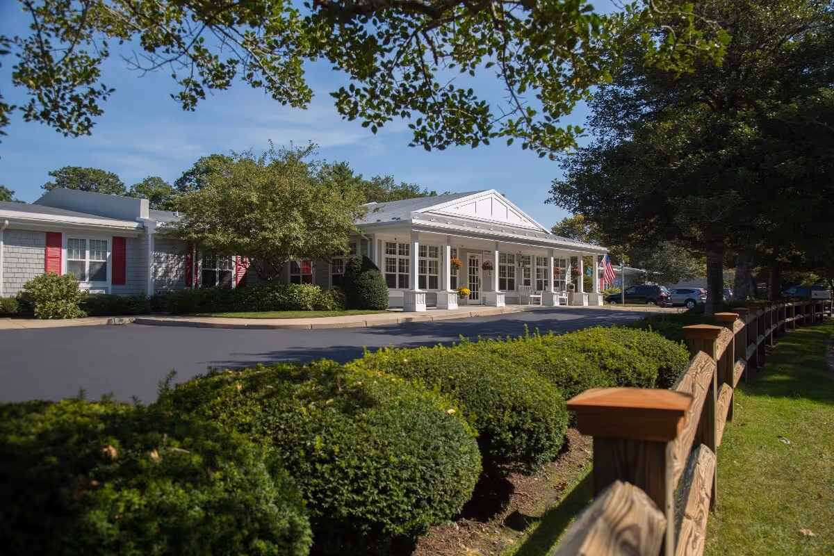 Exterior view of Harbor Point at Centerville, showing a single-story building with a white porch, columns, and hanging flower baskets. The building is surrounded by green bushes, trees, and a wooden fence along a grassy area under a clear blue sky.