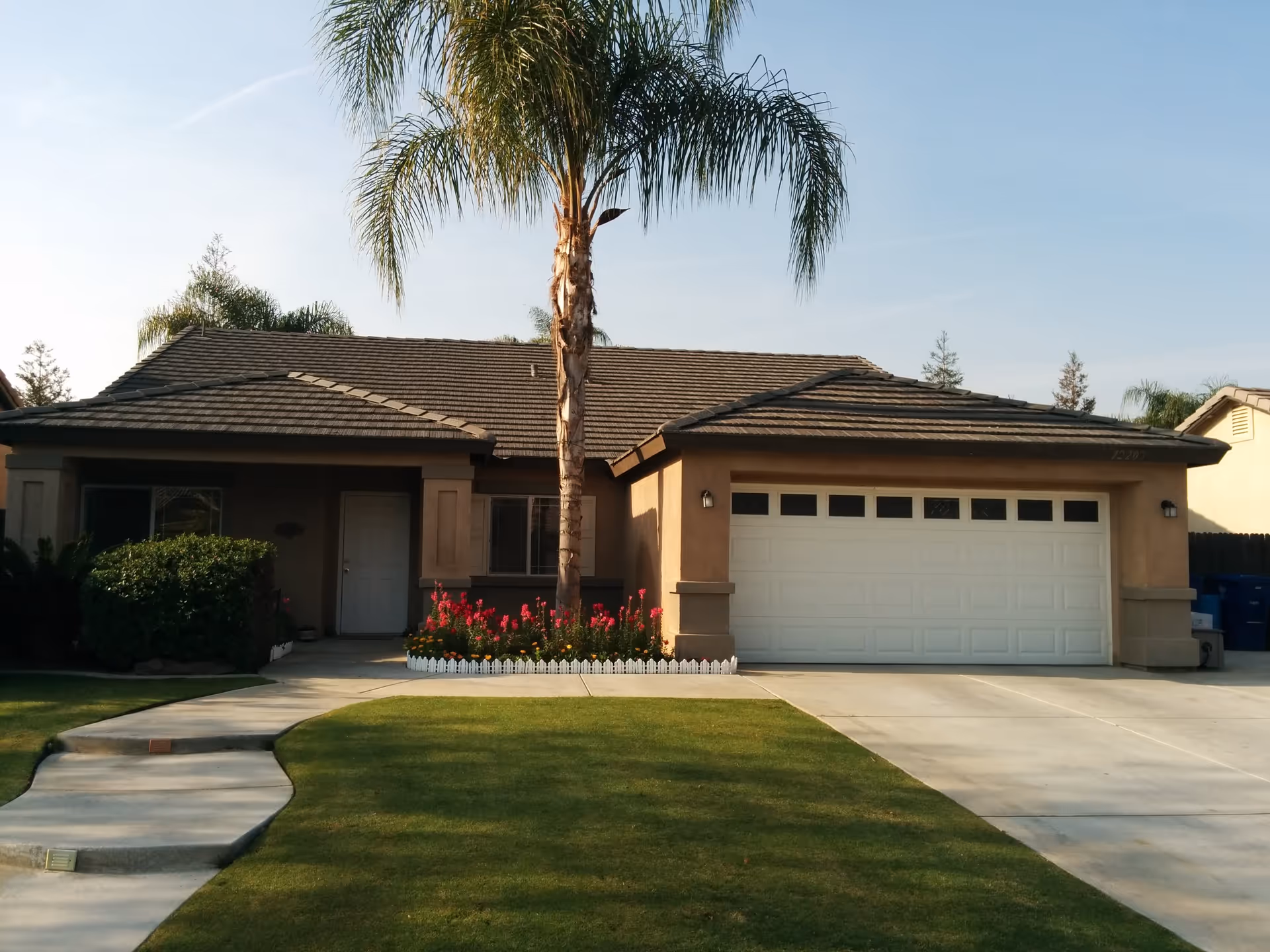 Single-story house with a tiled roof, a large palm tree in front, a white garage door, a small garden with red flowers bordered by a white picket fence, and a concrete driveway and walkway leading to the front door.