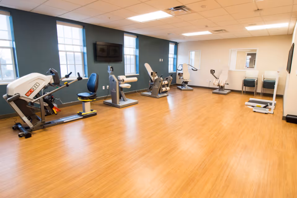 A spacious exercise room with wooden flooring and several pieces of fitness equipment arranged along the walls. The room has multiple windows letting in natural light, a wall-mounted TV, and a stack of chairs in the corner.