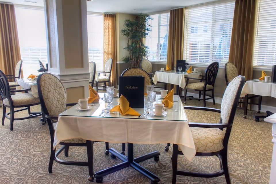 A bright dining room with several tables covered in white tablecloths and set with cups, glasses, silverware, and yellow folded napkins. The chairs have patterned upholstery and dark wooden frames. Large windows with blinds and mustard-colored curtains allow natural light to fill the room. A menu labeled 'Peakview' is placed on the tables.
