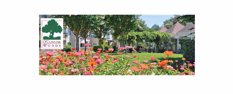 A vibrant garden with colorful flowers in the foreground, green grass, trees, and a pergola-covered seating area in the background at O'Connor Woods facility.