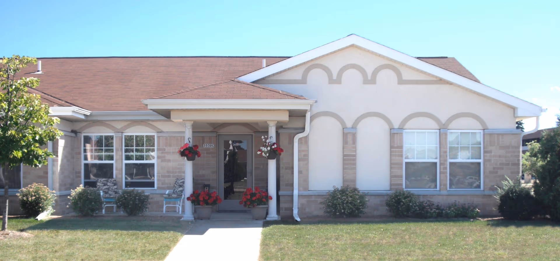 Front exterior of a one-story senior living building showing a covered entrance with columns, potted flowers, and multiple windows.