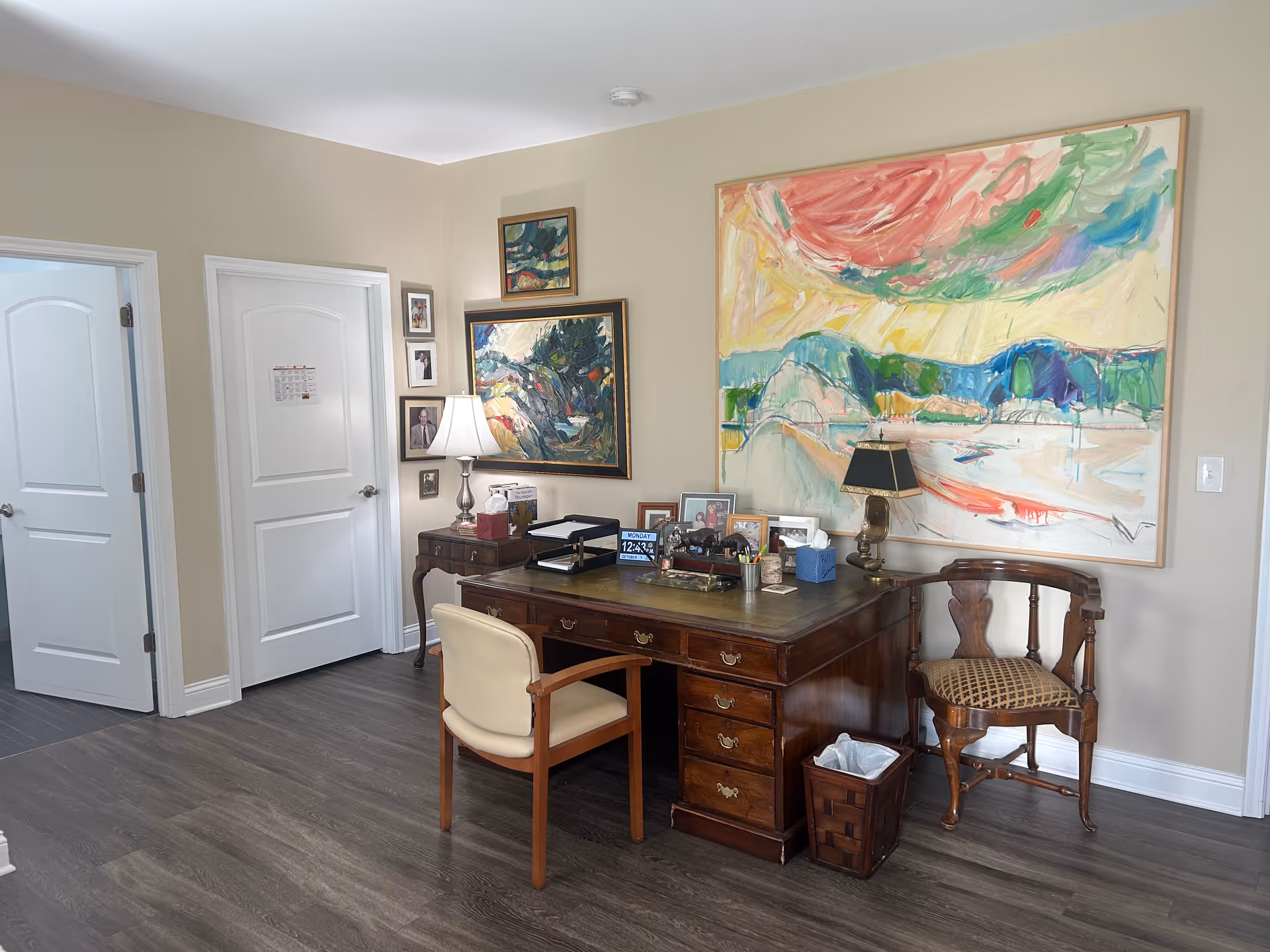 An office area in a senior living facility with a wooden desk and two chairs. The desk holds various items including framed photos, a lamp, and office supplies. The walls are decorated with colorful abstract paintings and several framed photographs. There are two white doors on the left side of the room and the floor is covered with dark wood laminate.