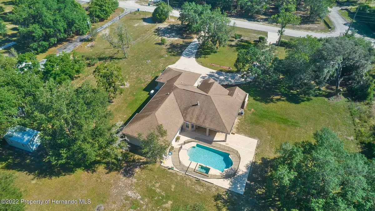 Aerial view of a single-story building with a brown roof surrounded by green trees and grass. The building has a fenced swimming pool and a hot tub in the backyard. A driveway leads from the building to a nearby road.