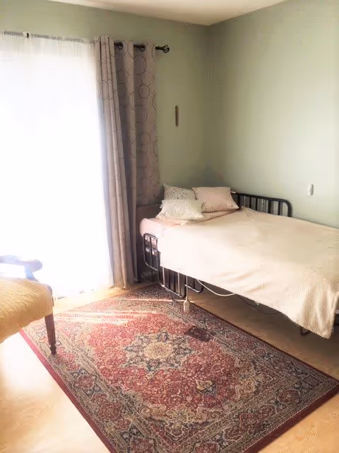 A small bedroom with a single metal-framed bed covered with a light beige blanket and several pillows. Next to the bed is a window with sheer white curtains and patterned gray drapes. A patterned area rug covers part of the wooden floor, and a wooden chair is partially visible on the left side.