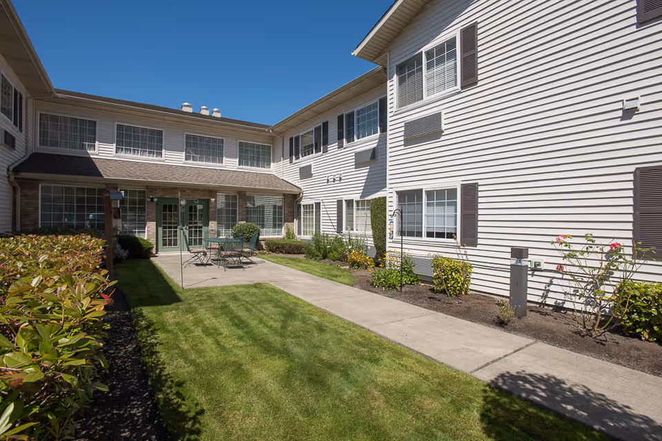 Sunlit courtyard with a walkway, lawn, patio table and chairs, and a two-story white-siding building with multiple windows.