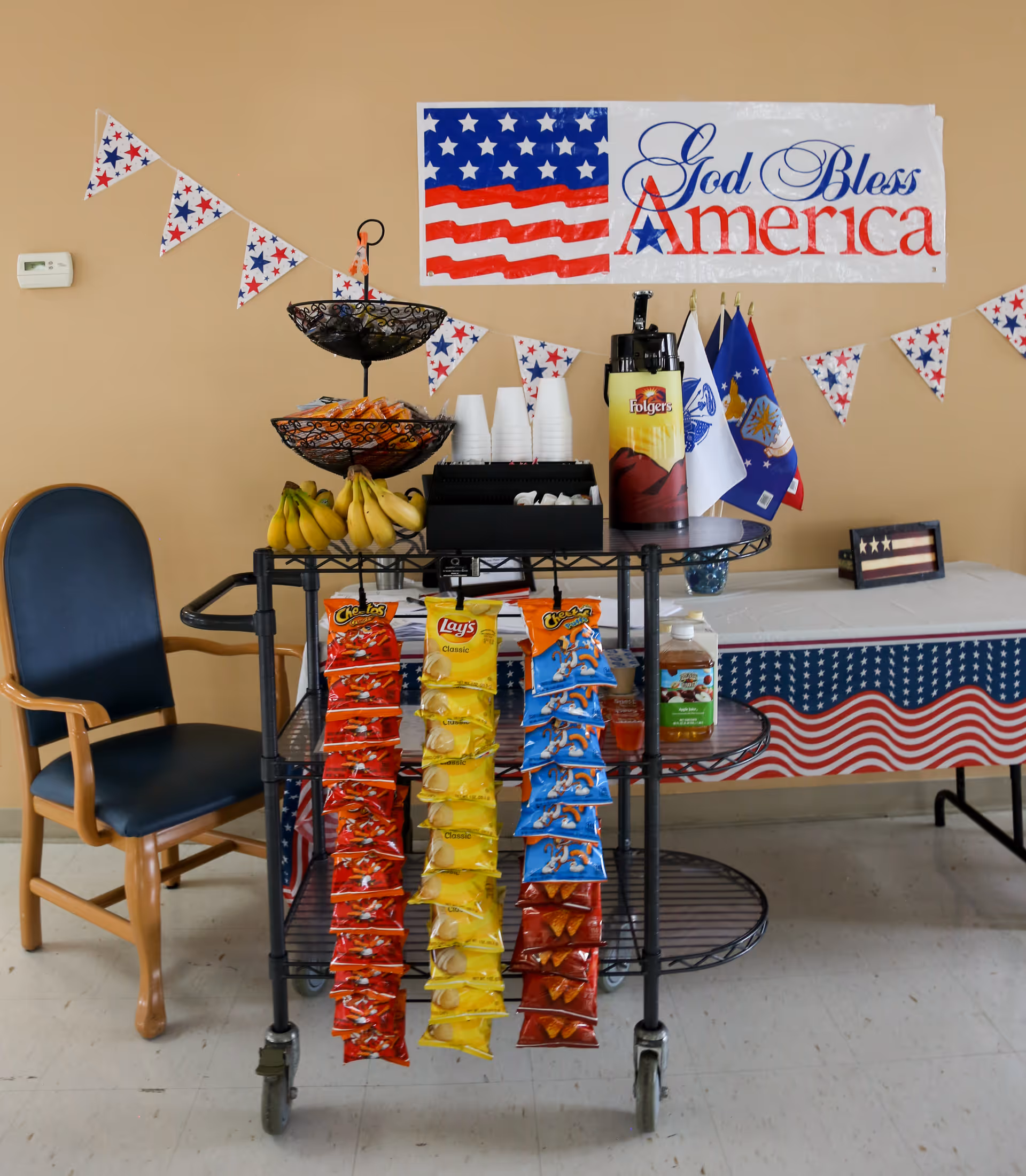 A snack cart decorated with patriotic red, white, and blue bunting and a 'God Bless America' banner on the wall behind it. The cart holds bananas, various bags of chips, a Folgers coffee dispenser, disposable cups, and small flags. A wooden chair with blue upholstery is positioned to the left of the cart, and a table covered with a patriotic tablecloth is in the background.