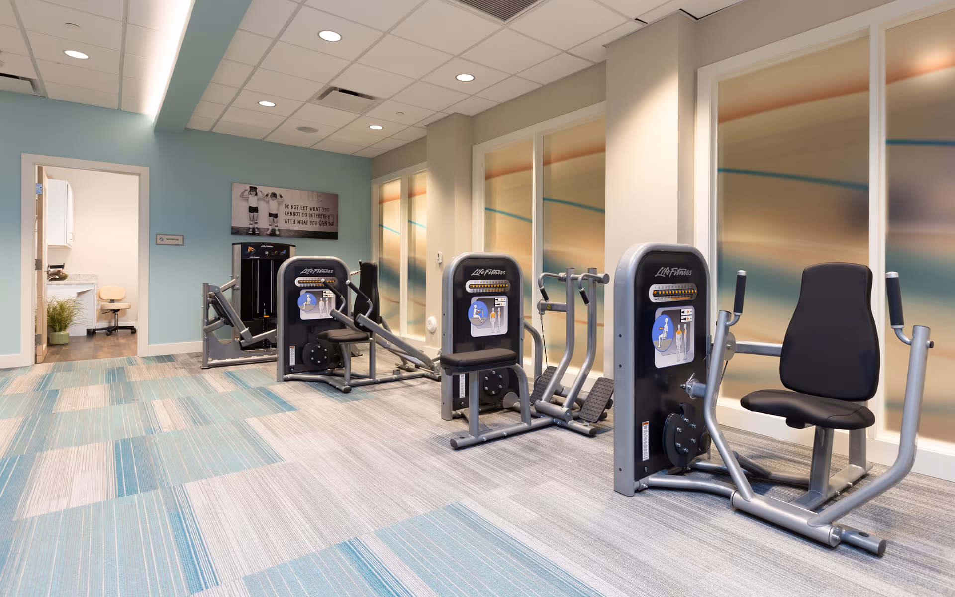 A fitness room with three Life Fitness exercise machines arranged in a row on a carpeted floor with blue and gray patterns. The walls are painted light blue and beige, with frosted glass panels featuring abstract colored lines. There is an open door leading to a small room with a chair and countertop. A motivational sign is mounted on the wall above the machines.