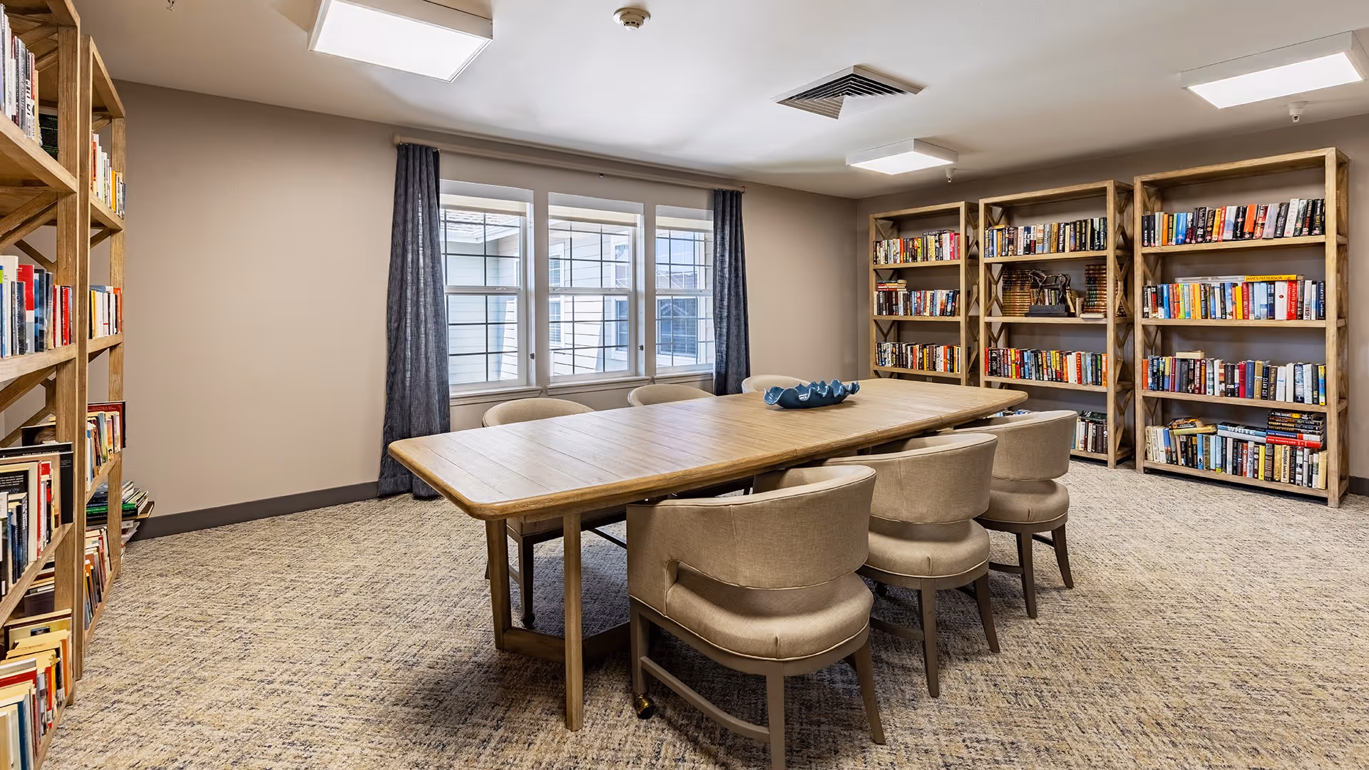A cozy library or reading room with wooden bookshelves filled with books along the walls, a large wooden table in the center surrounded by eight cushioned chairs, and a window with dark curtains letting in natural light.
