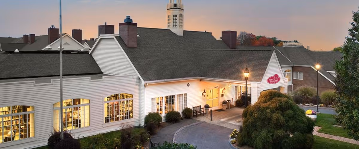 Exterior view of Avery Heights senior living facility at dusk, showing a white building with large windows illuminated from inside, a driveway with benches and street lamps, and surrounding greenery.