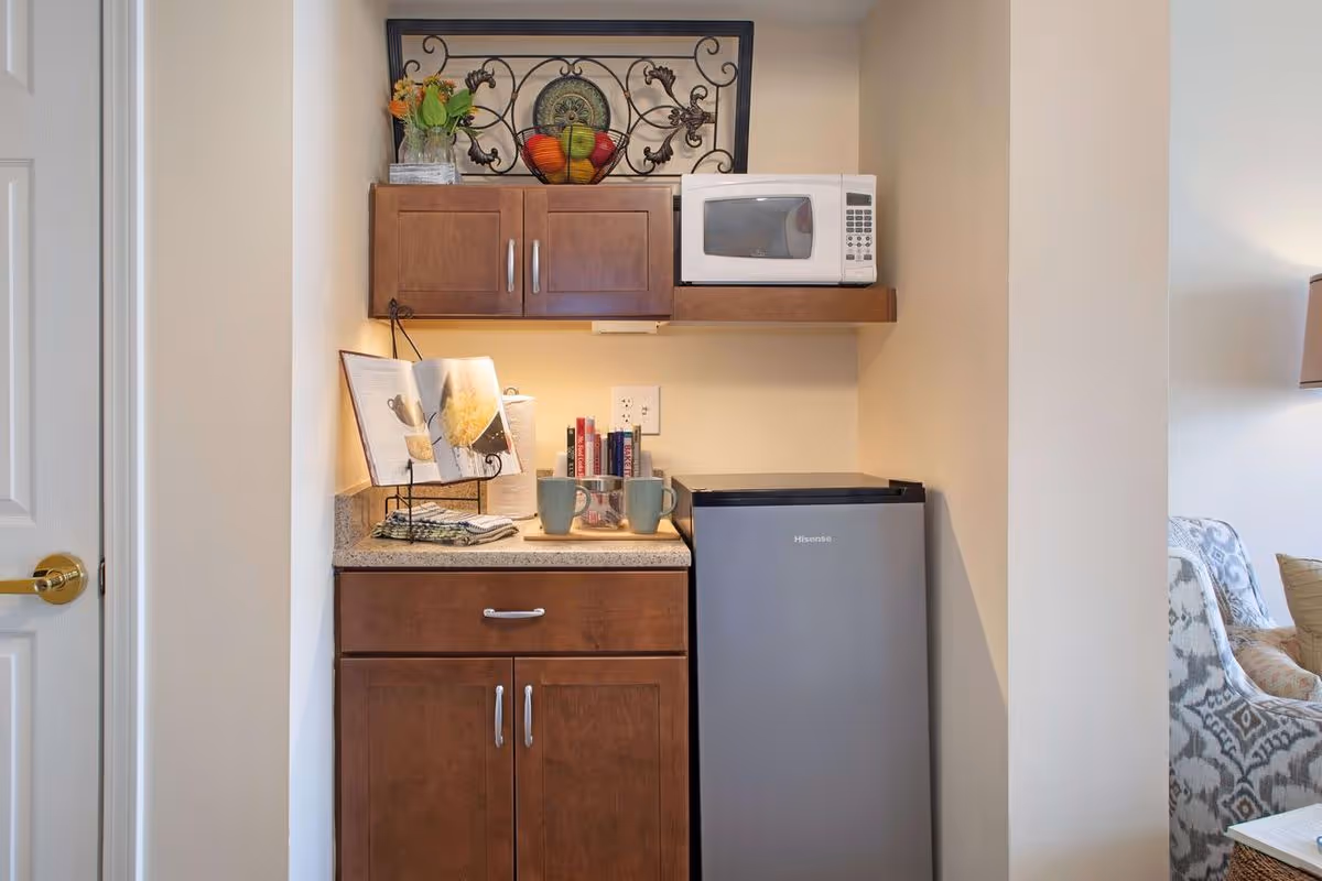 A small kitchenette area with wooden cabinets, a granite countertop, a mini refrigerator, and a microwave mounted on a wooden shelf. On the countertop, there are two mugs, a cookbook on a stand, some books, and a small vase with flowers. Part of a living room with a patterned armchair and a lamp is visible to the right.