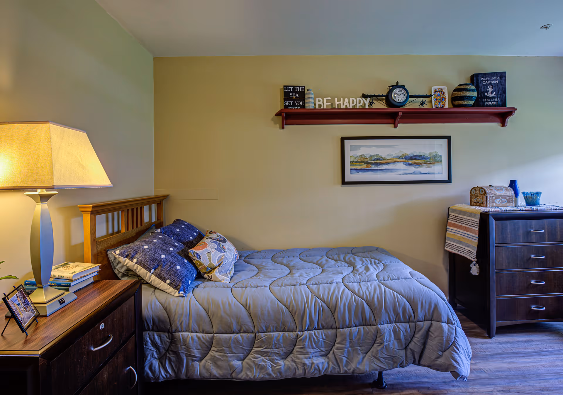 A neatly made single bed in a bedroom with a bedside lamp, nightstand, dresser, and a decorative shelf and framed artwork on the wall.