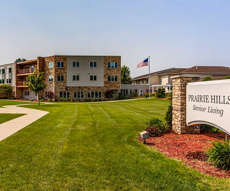 Exterior view of Prairie Hills Senior Living facility showing a multi-story building with stone and siding facade, a well-maintained green lawn, a sidewalk, an American flag on a flagpole, and a sign that reads 'Prairie Hills Senior Living'.