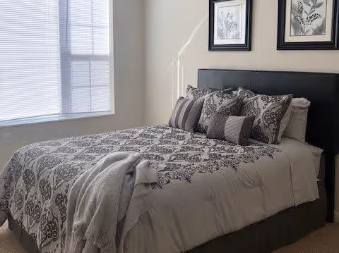 A neatly made bed with patterned bedding and multiple pillows in a bedroom with a window covered by blinds and two framed botanical prints on the wall above the headboard.