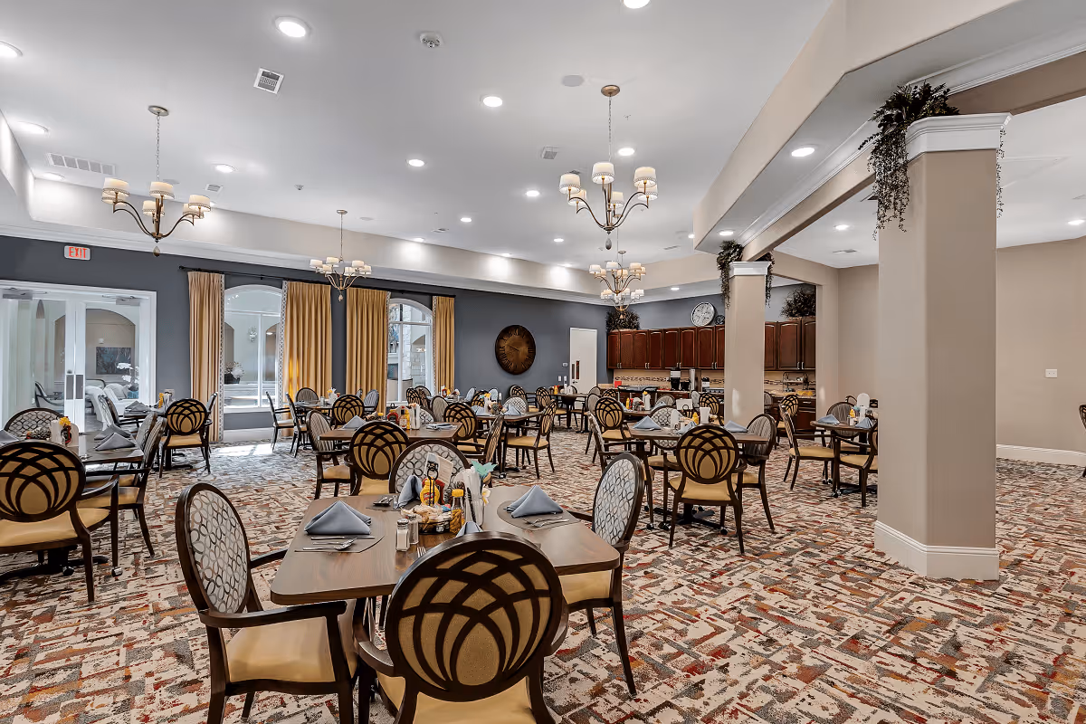 A spacious dining room with multiple tables and chairs arranged neatly. The room features patterned carpet, beige walls, and large windows with yellow curtains allowing natural light to enter. There are chandeliers hanging from the ceiling and a kitchen area with wooden cabinets in the background.