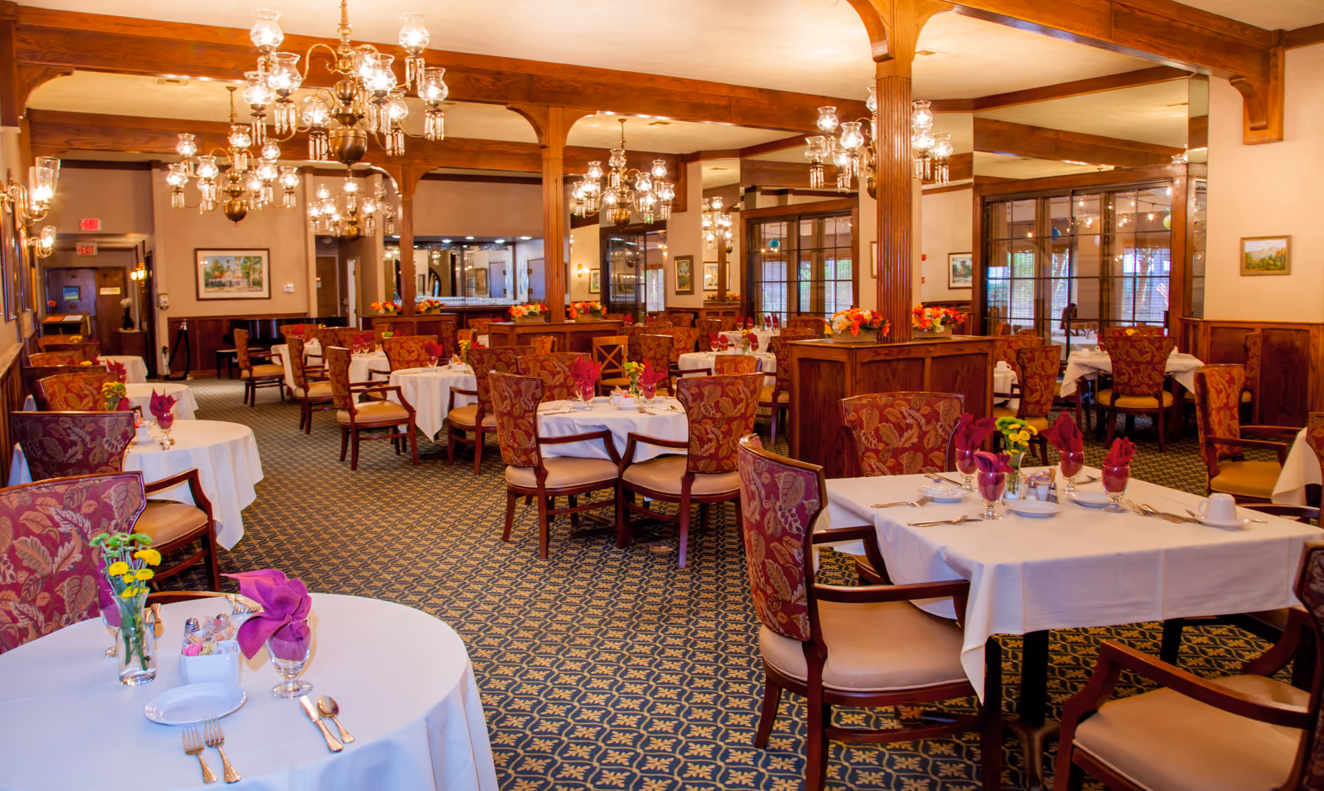 A spacious dining room with multiple round and square tables covered with white tablecloths. Each table is set with silverware, glasses with folded purple napkins, and small flower arrangements. The room features patterned carpet, wooden columns, and ornate chandeliers hanging from the ceiling. The walls have framed artwork and large windows with wooden frames.