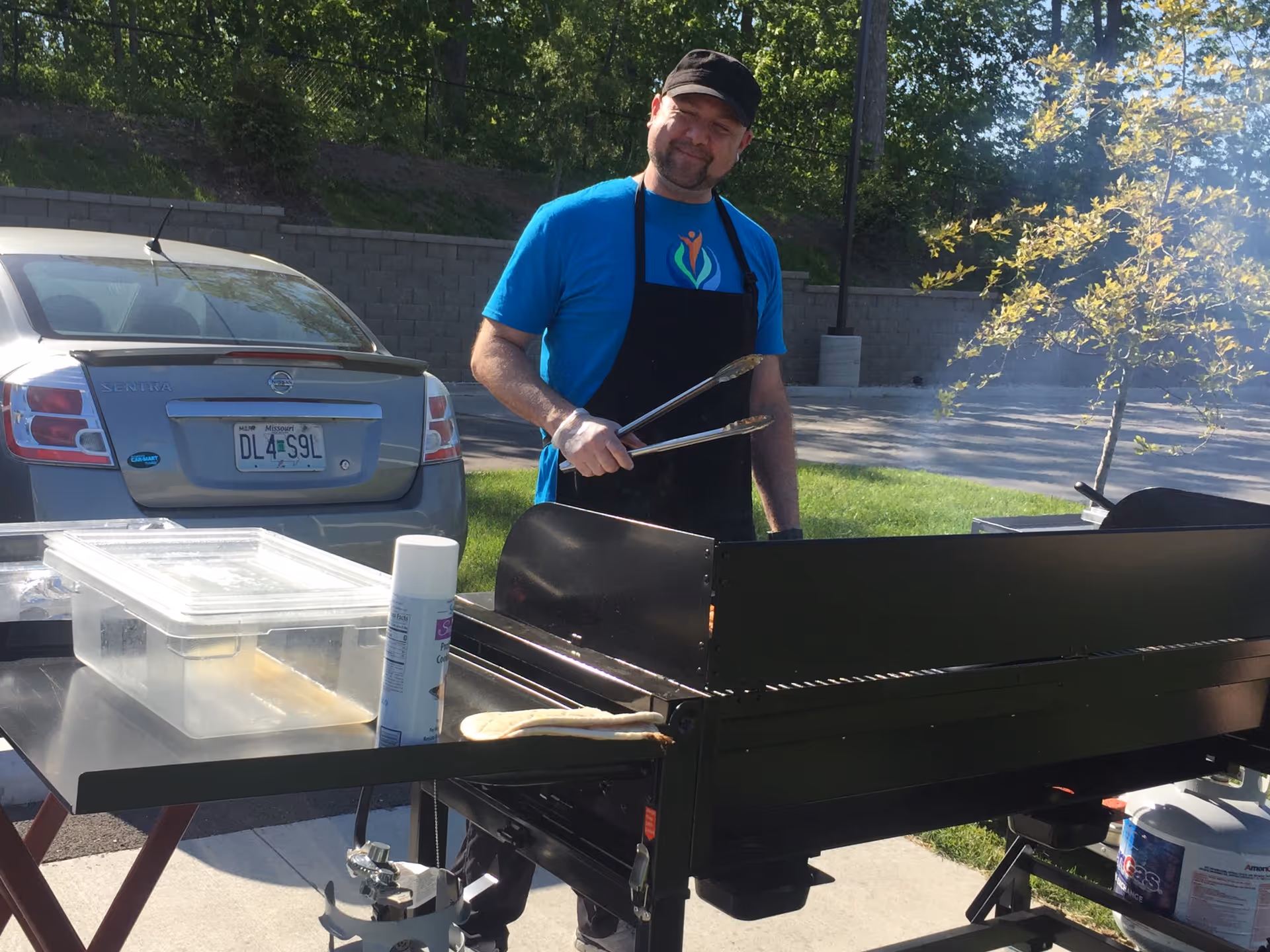 A man wearing a black cap, blue t-shirt, and black apron is standing outdoors next to a large black grill holding a pair of tongs. There is a silver car parked nearby and some green grass and trees in the background. The scene appears to be a sunny day.