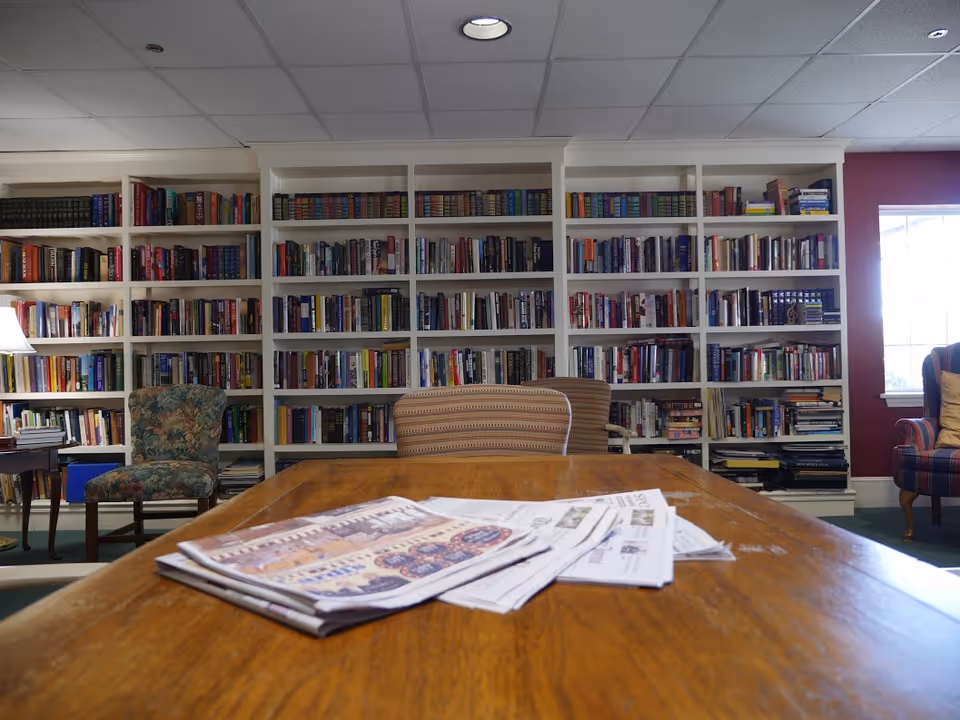 A cozy reading room with a large wooden table in the foreground holding newspapers, surrounded by several upholstered chairs. Behind the chairs is a wall filled with white bookshelves packed with books. A window on the right side lets in natural light.