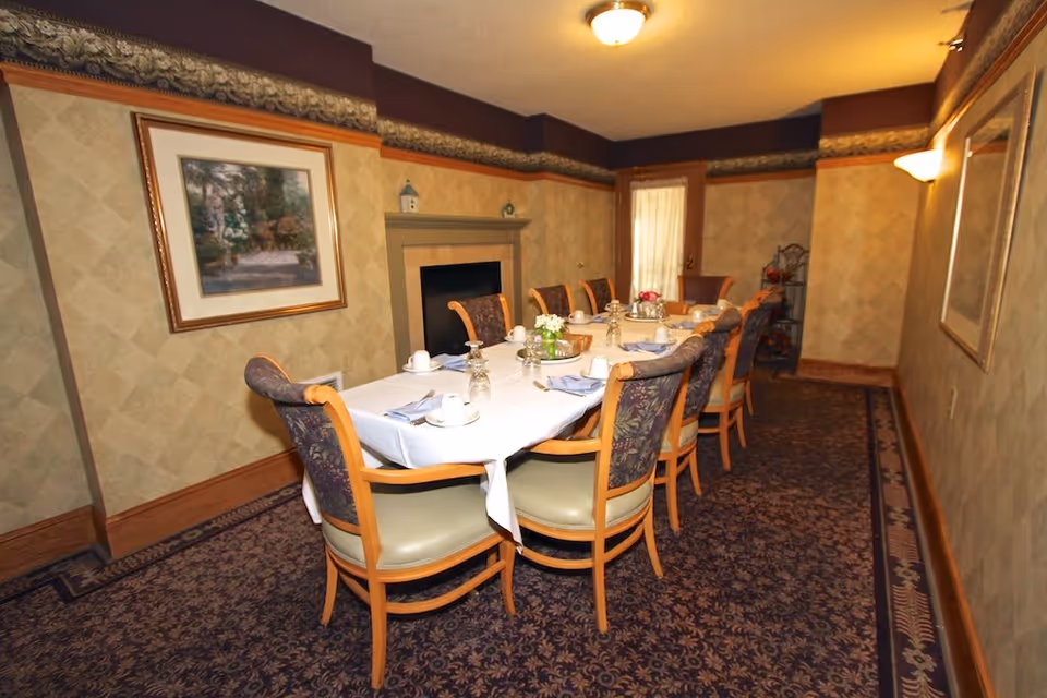 A dining room with a long rectangular table covered with a white tablecloth, set with cups, saucers, napkins, and small flower arrangements. The room has patterned carpet, wallpaper with a decorative border near the ceiling, a framed painting on the wall, a fireplace, and a door with a sheer curtain.