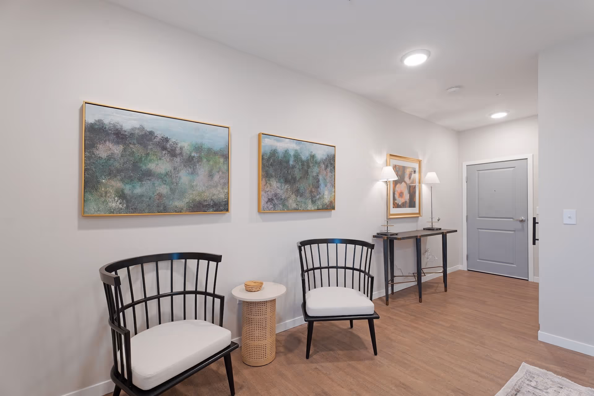 A hallway area in a senior living facility with two black chairs with white cushions, a small round side table between them, and a console table with two lamps and a framed floral picture above it. Two abstract landscape paintings hang on the wall above the chairs. The floor is wood, and there is a gray door at the end of the hallway.