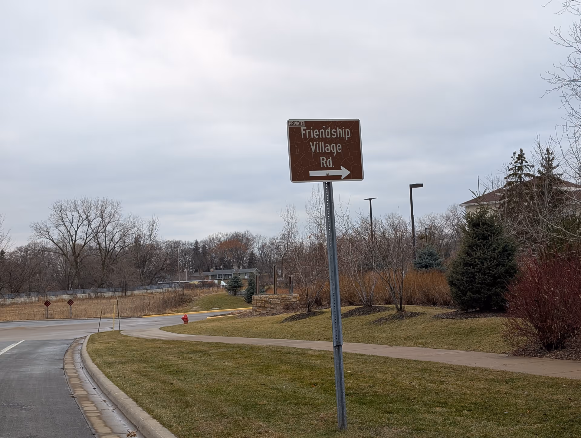 A brown road sign on a metal pole beside a curved road and sidewalk, pointing right and labeled 'Friendship Village Rd.' with the word 'PRIVATE' in the top left corner of the sign. The scene includes grass, leafless trees, some evergreen shrubs, and a cloudy sky.