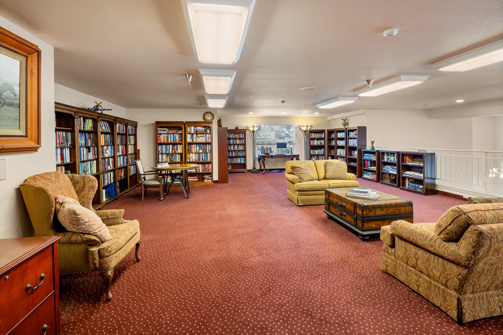 A spacious senior living facility library with red carpet flooring and beige walls. The room features multiple wooden bookshelves filled with books along the walls, a few comfortable upholstered armchairs and a loveseat with patterned cushions, a wooden coffee table in the center, and a small table with chairs near the bookshelves. Large fluorescent ceiling lights illuminate the room, and a window at the far end lets in natural light.