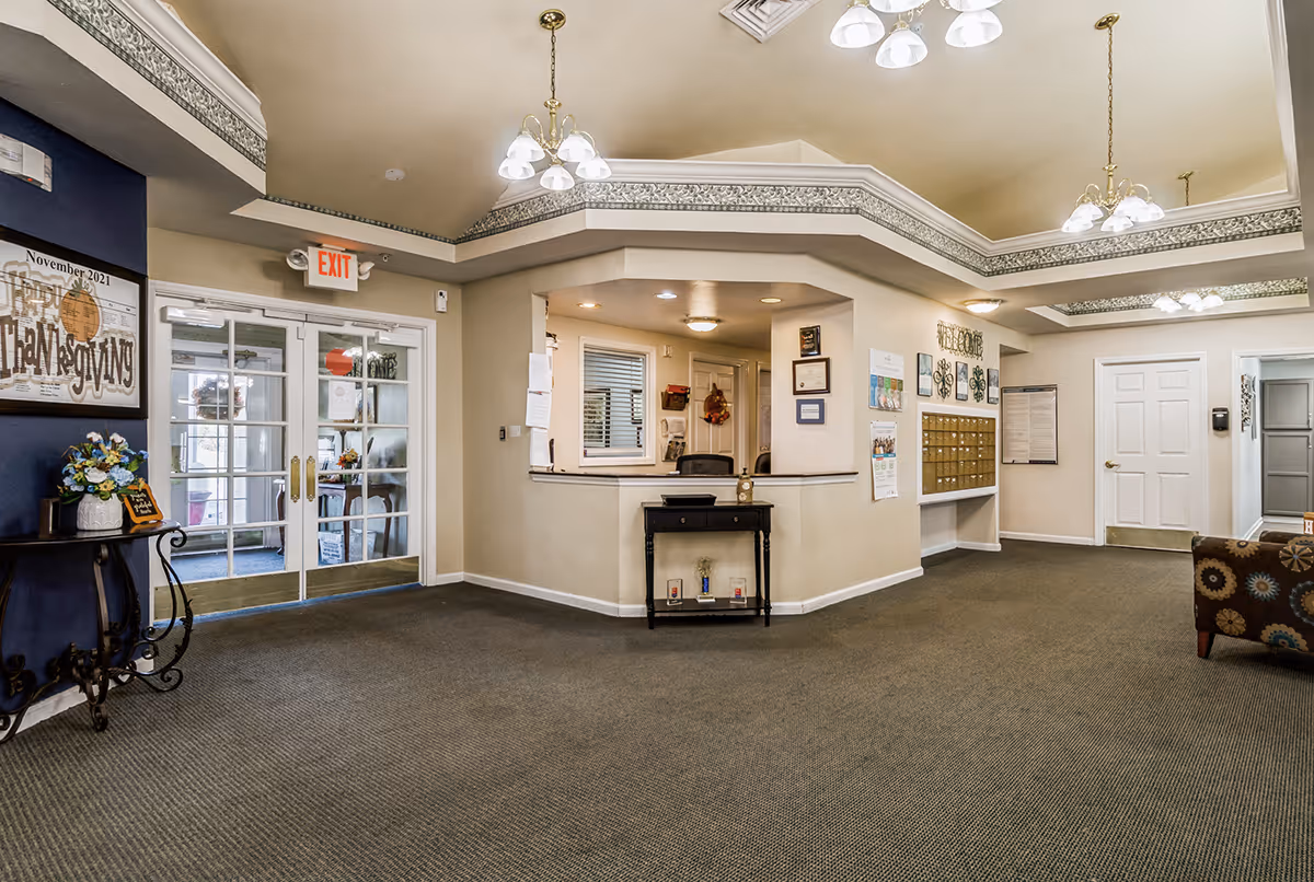Interior view of a senior living facility lobby area with a reception desk, carpeted floor, decorative table with flowers, mailboxes on the wall, and double glass doors leading outside. The ceiling has crown molding and hanging light fixtures.