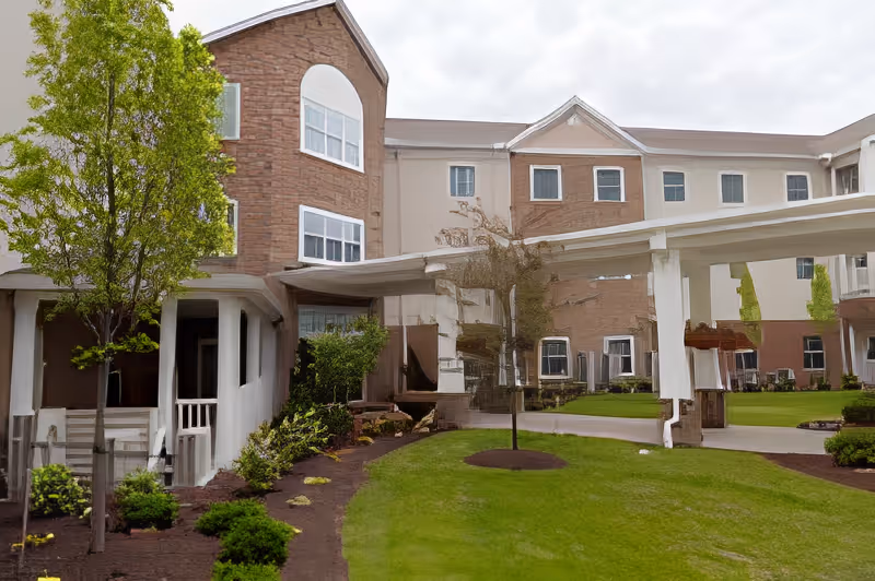Exterior view of Sanctuary Grande senior living facility showing a multi-story building with brick and beige walls, several windows, a covered walkway, and landscaped green lawn with trees and shrubs.