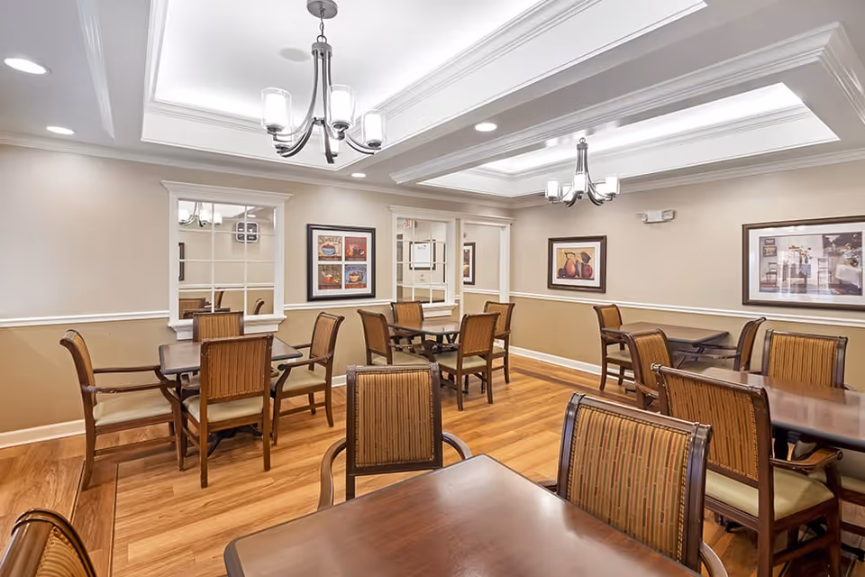 A well-lit dining room with multiple wooden tables and cushioned chairs arranged neatly. The room features light beige walls with white trim, framed artwork on the walls, and two chandeliers hanging from a coffered ceiling. The floor is made of wood, and there are windows with white frames on one wall.