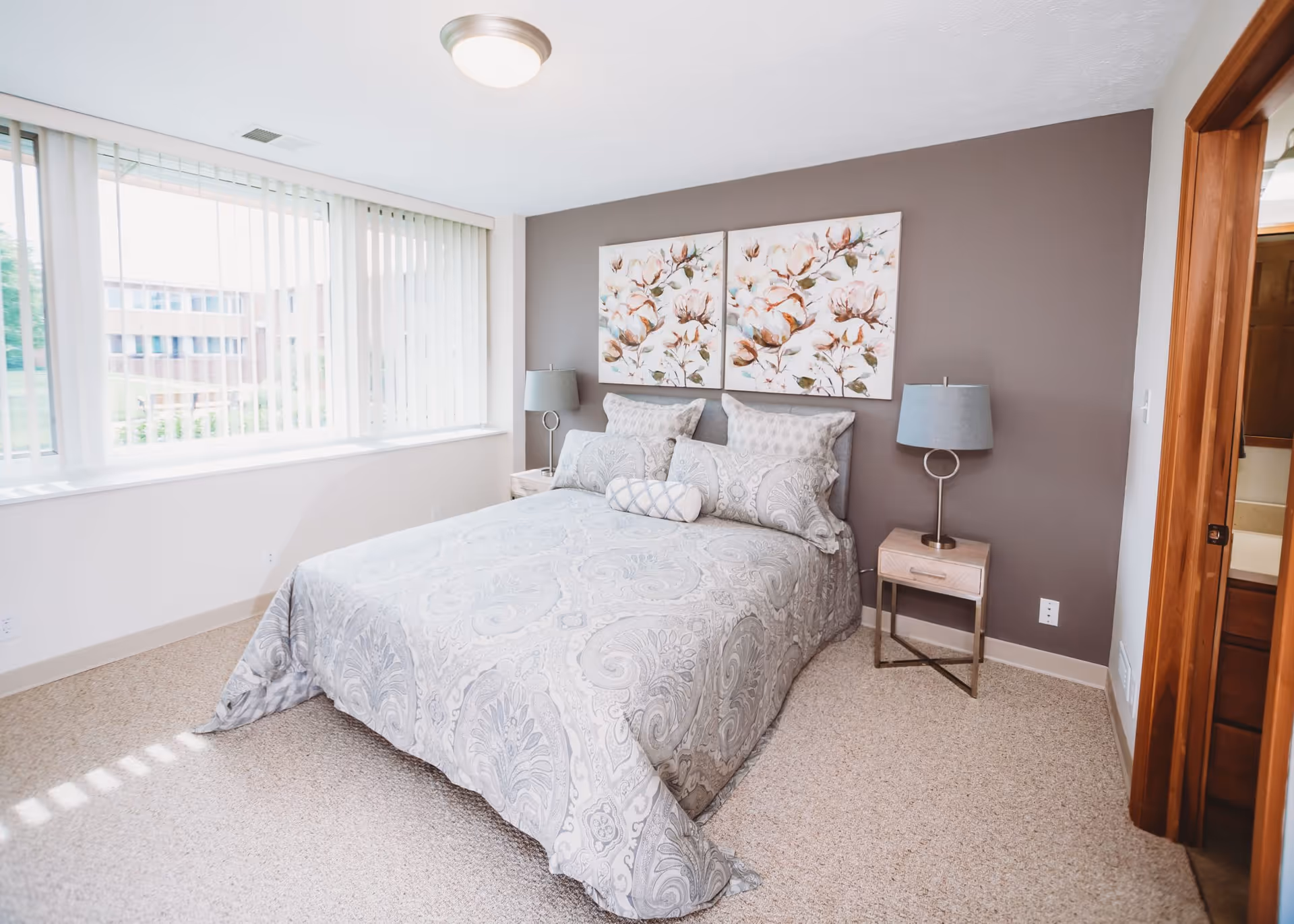 A bright bedroom with a large window covered by vertical blinds, a bed with patterned gray bedding and multiple pillows, two bedside tables each with a lamp, and two floral paintings on a gray accent wall behind the bed.