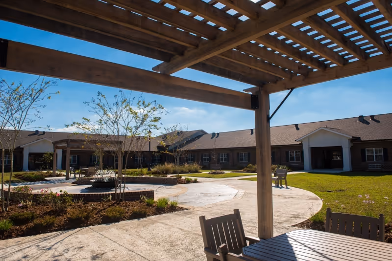 Sunlit courtyard with a wooden pergola, outdoor seating, a central fountain, and surrounding single-story brick buildings.