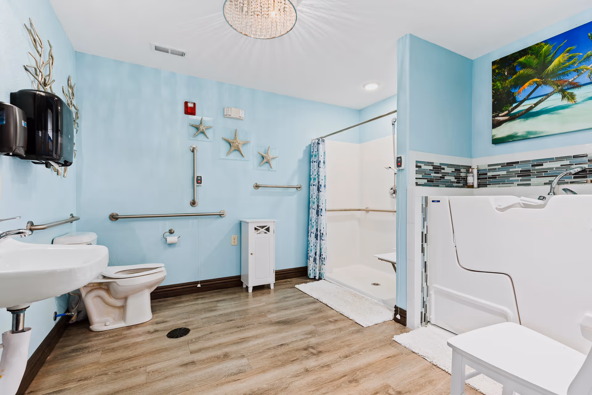 A spacious, accessible bathroom with light blue walls and wood-look flooring. The room features a white toilet, a wall-mounted sink, grab bars, a walk-in shower with a curtain, and a walk-in bathtub. There is a small white cabinet and a white chair in the room. The walls are decorated with starfish ornaments and a large tropical beach painting. Two black soap dispensers are mounted on the wall near the sink.