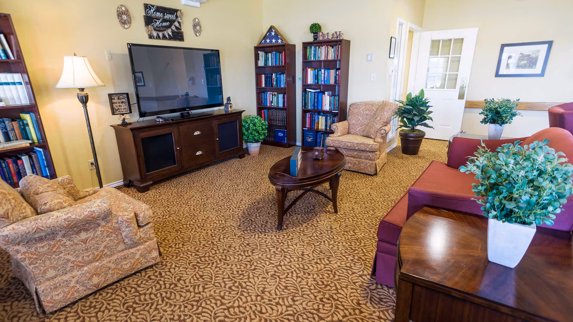 Comfortable communal living room with upholstered chairs and sofas, a flat-screen TV on a wooden console, bookshelves, a coffee table and potted plants.