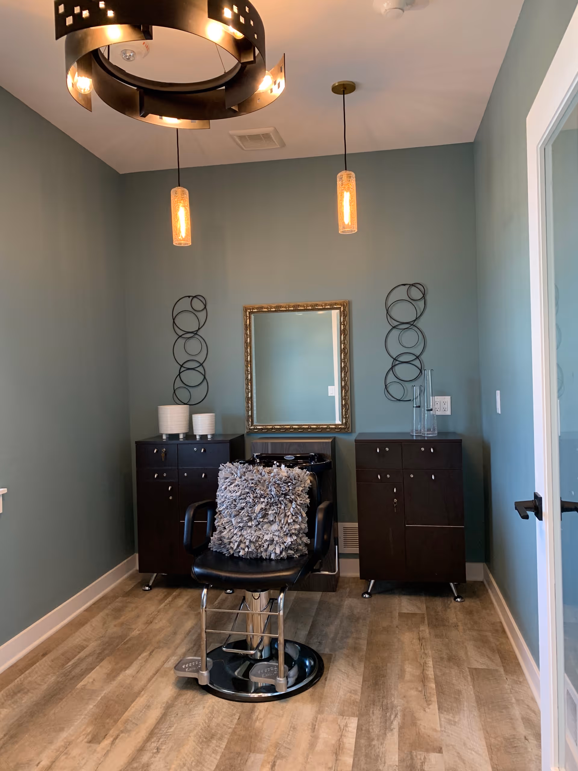 A small salon/styling room with a black barber chair in front of a framed mirror, flanked by two dark cabinets under pendant lights.
