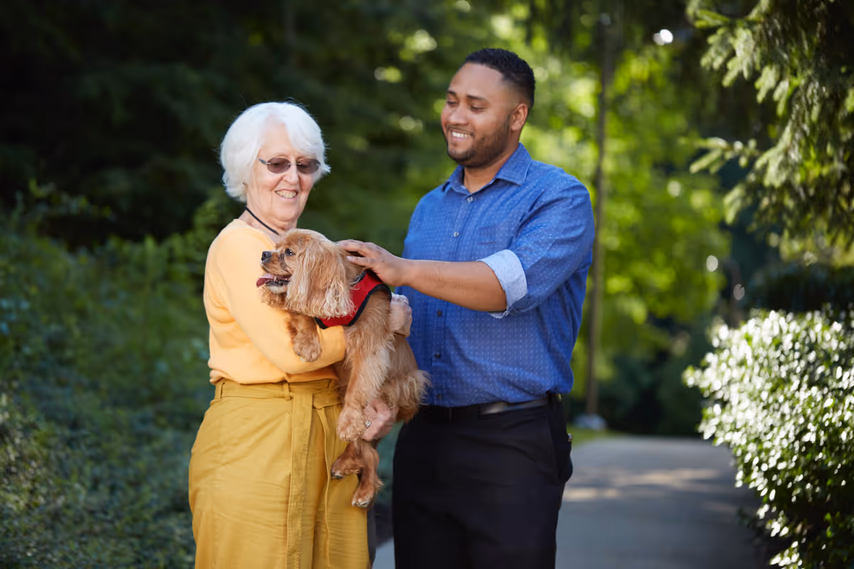 An elderly woman wearing glasses and a yellow outfit holds a small brown dog wearing a red harness. A man in a blue shirt stands next to her, smiling and petting the dog. They are outdoors on a path surrounded by greenery.