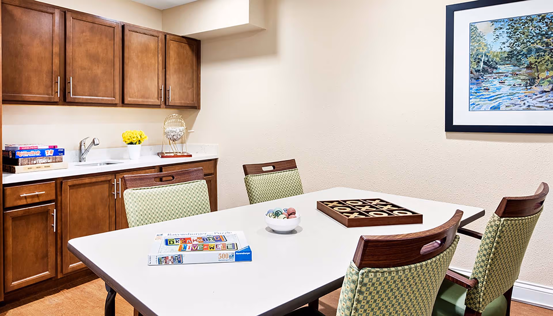 A game room with a white table surrounded by four green patterned chairs. On the table are a tic-tac-toe game, a bowl with bingo balls, and a puzzle box. Behind the table is a countertop with wooden cabinets above and below, a small sink, a yellow flower pot, and a bingo cage. A framed painting of a river scene hangs on the beige wall.