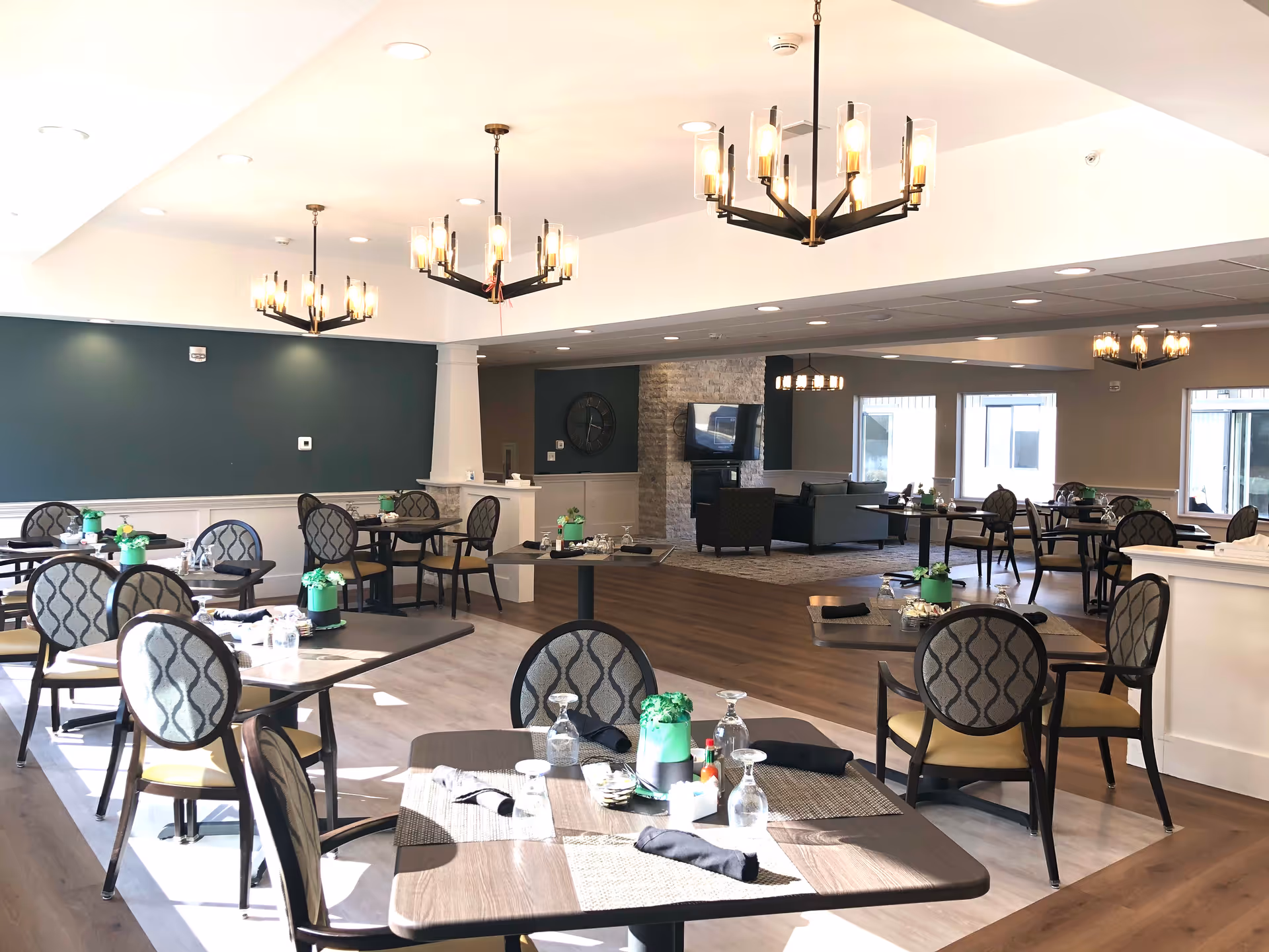 A bright and spacious dining area in a senior living facility with multiple tables and chairs arranged neatly. Each table is set with placemats, upside-down glasses, black napkins, and small green potted plants. The room features modern chandeliers hanging from the ceiling, a mix of light wood and darker flooring, and a cozy seating area with a TV and armchairs in the background.