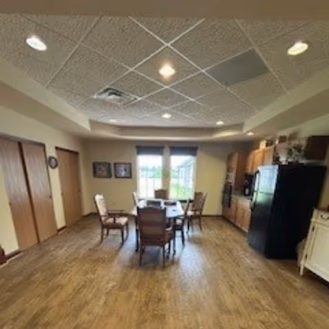 A dining area with a wooden table and six chairs in the center of the room. The room has wood flooring, a ceiling with recessed lighting, and a window with blinds letting in natural light. On the right side, there is a kitchen area with wooden cabinets, a black refrigerator, and a microwave. The left side has two closed wooden doors and framed pictures on the wall.
