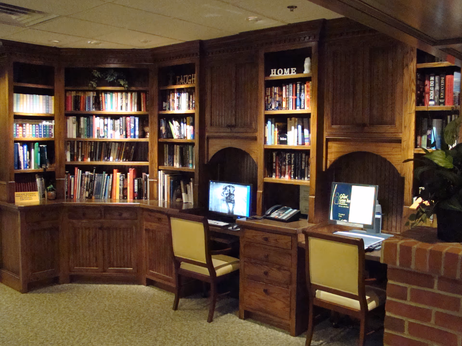 A cozy library or study area with wooden bookshelves filled with books. Two computer workstations with chairs are built into the shelves. The shelves have decorative words like 'LAUGH' and 'HOME'. There is a brick half-wall on the right side with a potted plant on top.