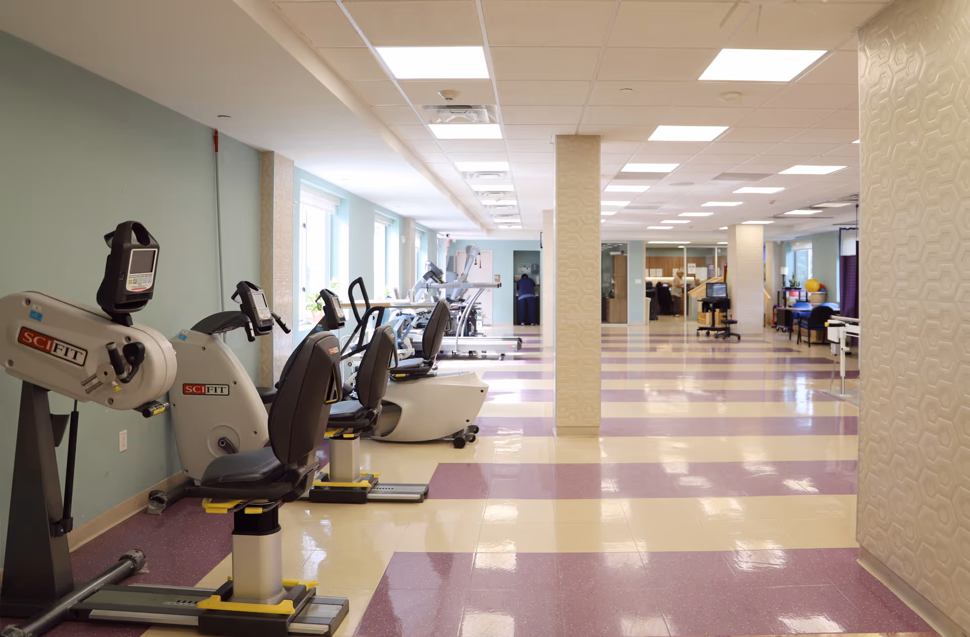 Spacious, well-lit rehabilitation gym with row of recumbent exercise bikes and cardio equipment on a purple-and-white tiled floor.