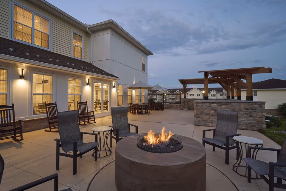 Outdoor patio area at dusk with a circular fire pit surrounded by chairs. The patio is adjacent to a two-story building with lit windows and rocking chairs along the wall. There are tables with umbrellas and a wooden pergola structure on the right side.