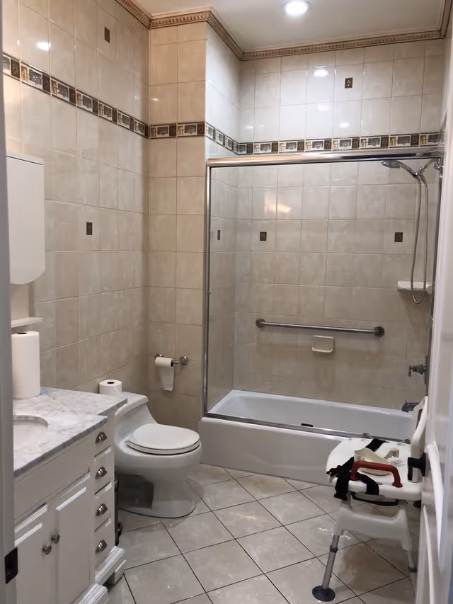 A clean bathroom with beige tiled walls and floor, featuring a white toilet, a bathtub with a sliding glass door and a showerhead, a white vanity with a marble countertop, and a white shower chair with red handles positioned near the bathtub.