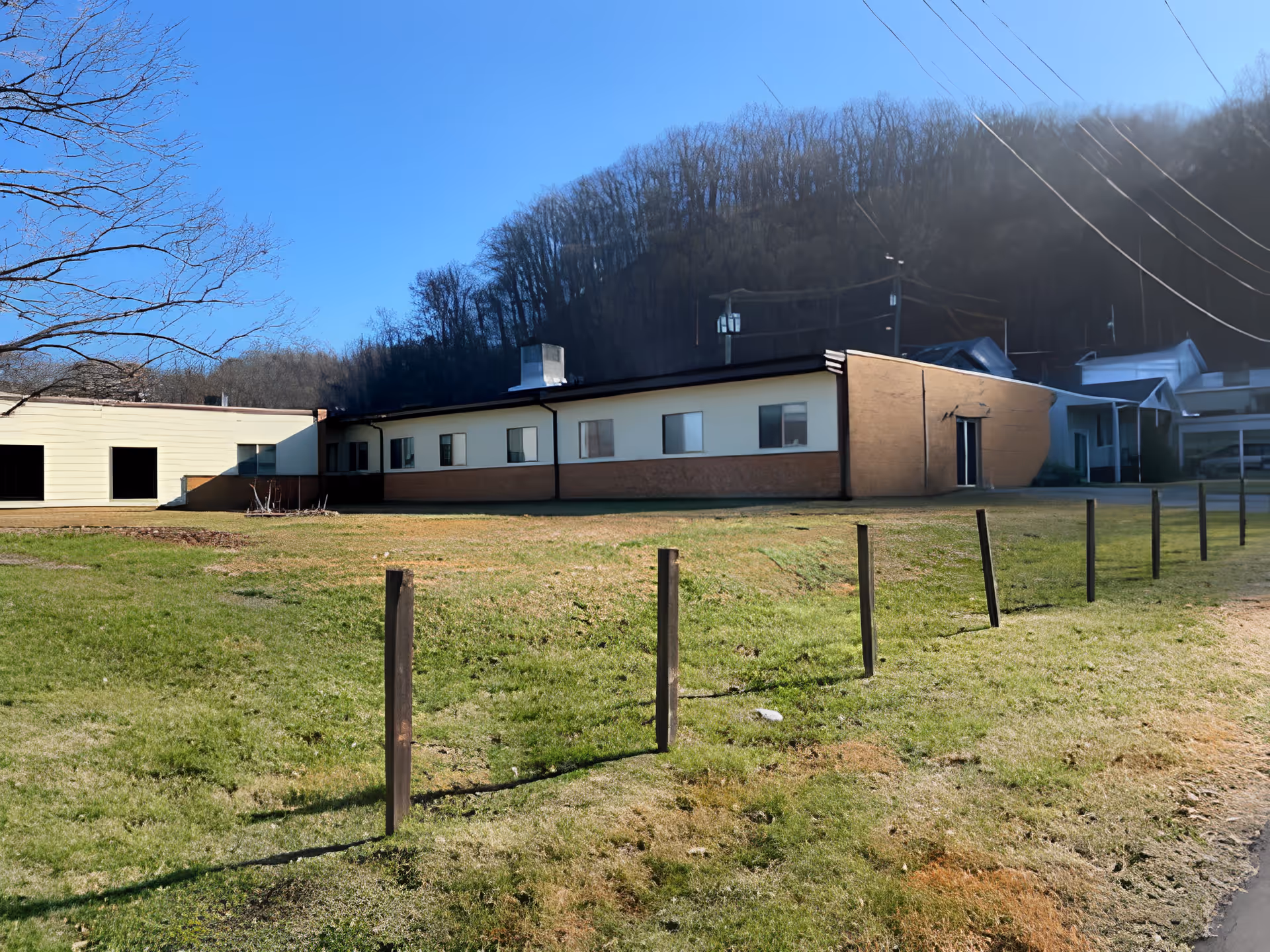 Single-story brick and siding building with a grassy lawn and a row of wooden posts in front, set against a wooded hillside under a clear blue sky.