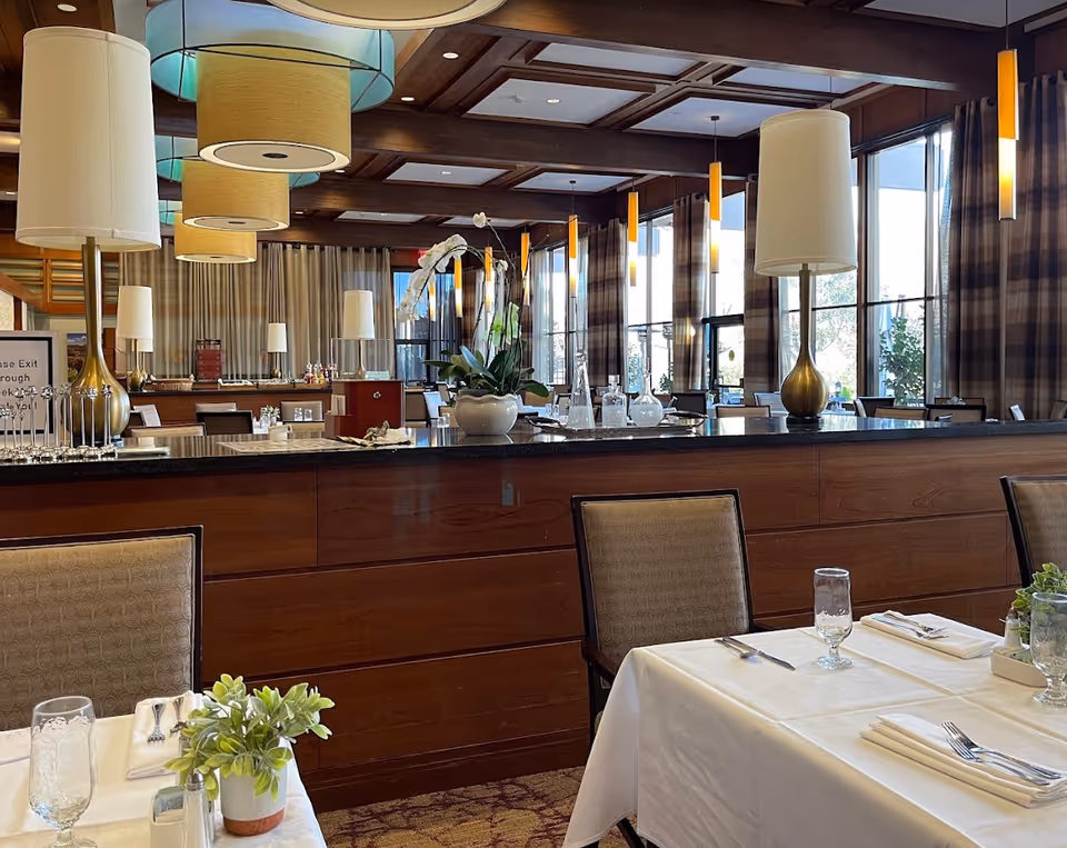 Upscale dining room with set tables, potted plants, large lamps, and a wooden service counter under pendant lighting.