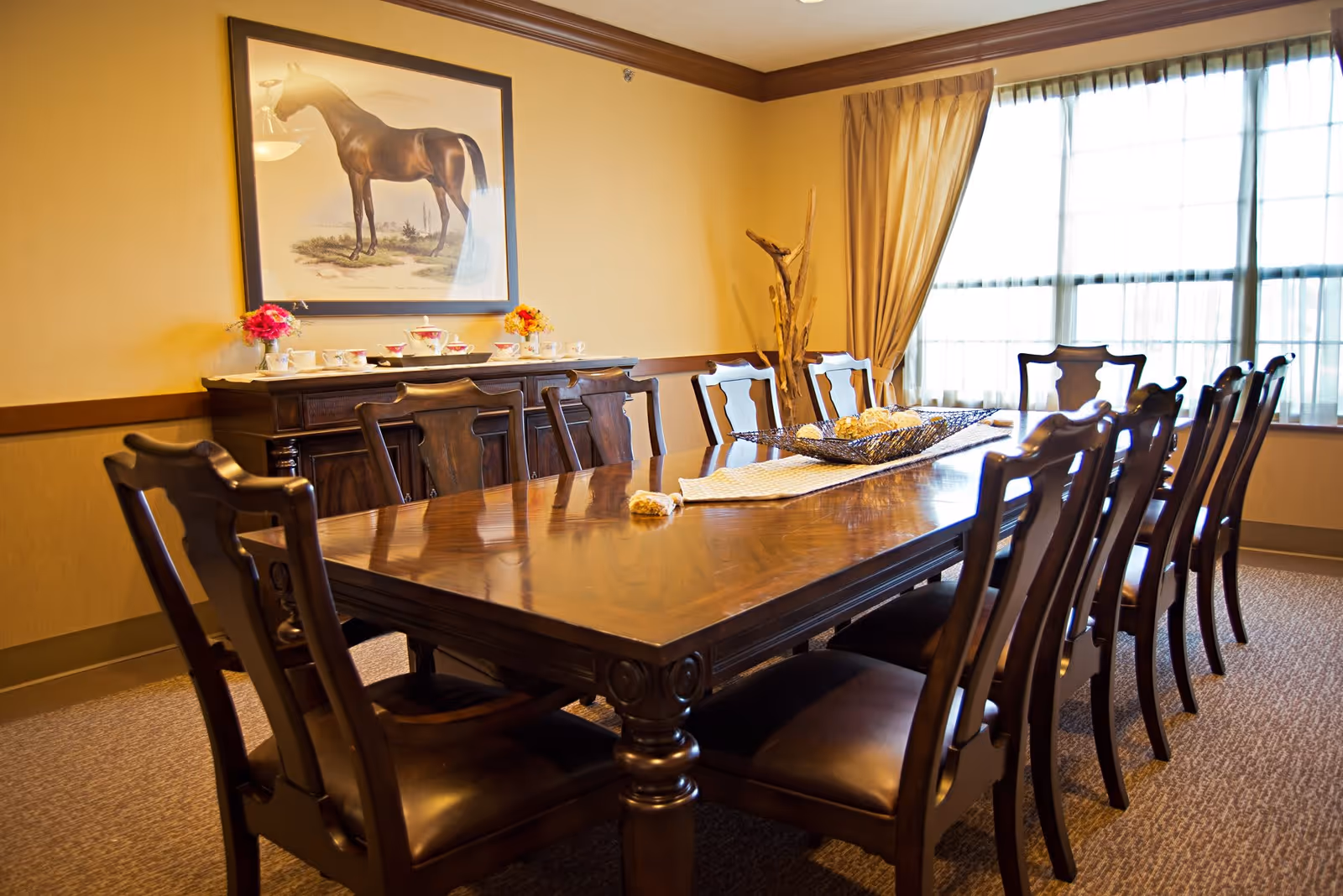 A formal dining room with a long polished wooden table surrounded by ten matching wooden chairs with leather seats. A sideboard against the wall holds a tea set and small flower arrangements. A large framed painting of a horse hangs above the sideboard. The room has beige walls, a large window with sheer curtains, and carpeted flooring.
