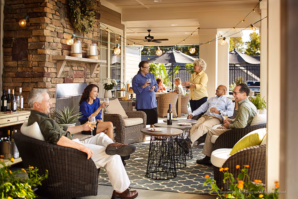 A group of older adults socializing and enjoying drinks on a covered outdoor patio with wicker furniture, a stone fireplace, string lights, and decorative plants.